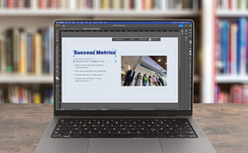 Macbook on table in front of bookshelves with InDesign on the screen