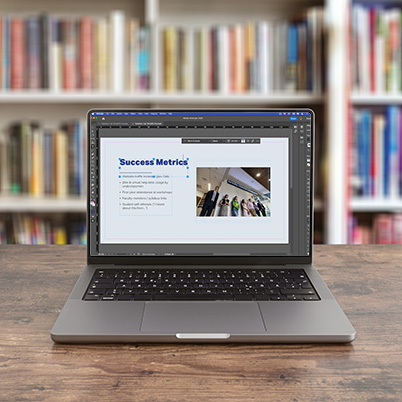 Macbook on table in front of bookshelves with InDesign on the screen